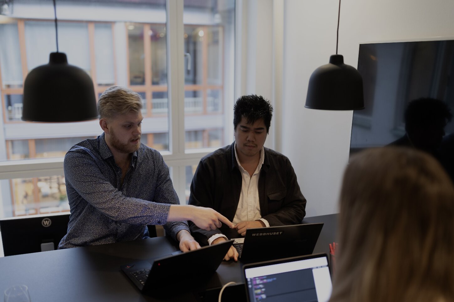 Två medarbetare sitter framför en laptop och arbetar tillsammans. En tredje person sitter mittemot med ryggen vänd från kameran och kollar på medarbetarna.